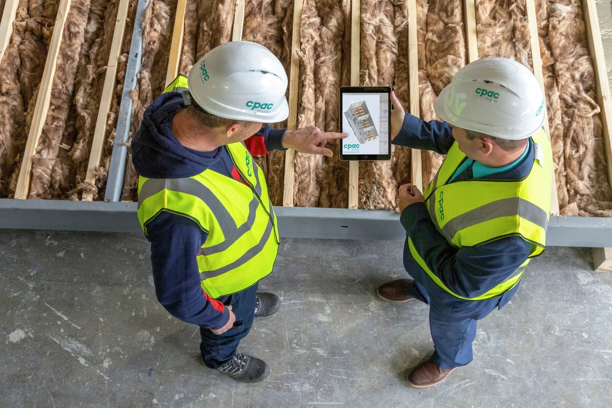 Zwei Handwerker mit Helm und Warnweste besprechen auf einer Holzbau-Baustelle Pläne auf einem Tablet.